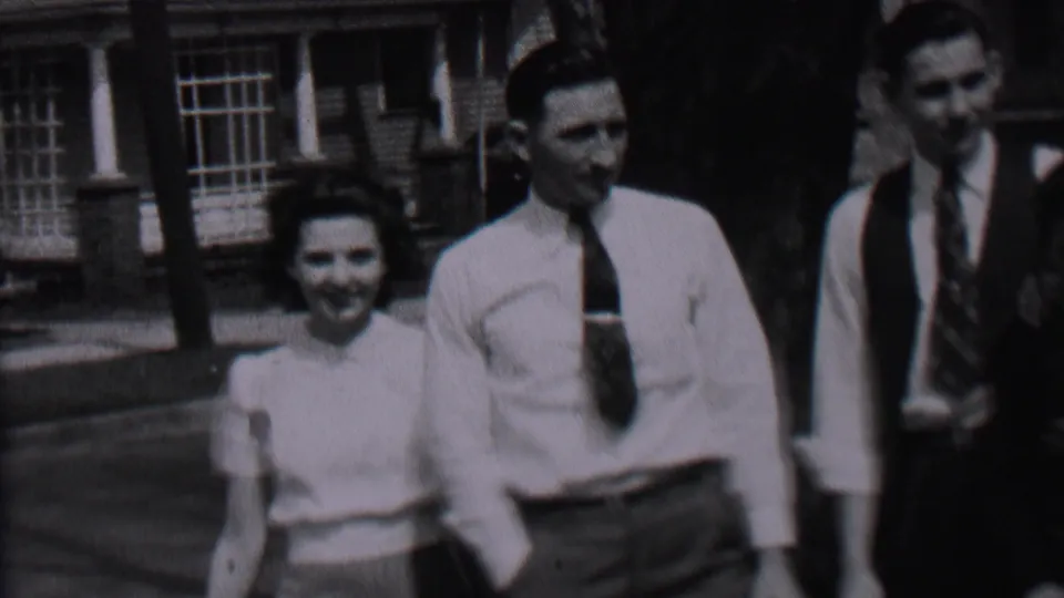 In 1937 Albany home-movie footage, a woman in a light blouse walks toward two men in shirts and ties near a brick residential-looking building.