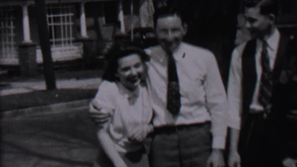 The woman leans close and smiles as a man in a white shirt puts an arm around her, with another suited or vested man visible nearby in the 1937 Albany home movie.
