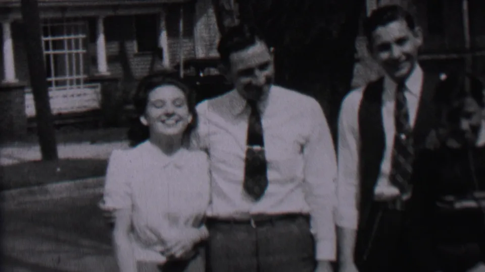 The woman smiles broadly while standing close to the man in a white shirt, with a second man in a vest and tie beside them in the Albany 1937 street footage.