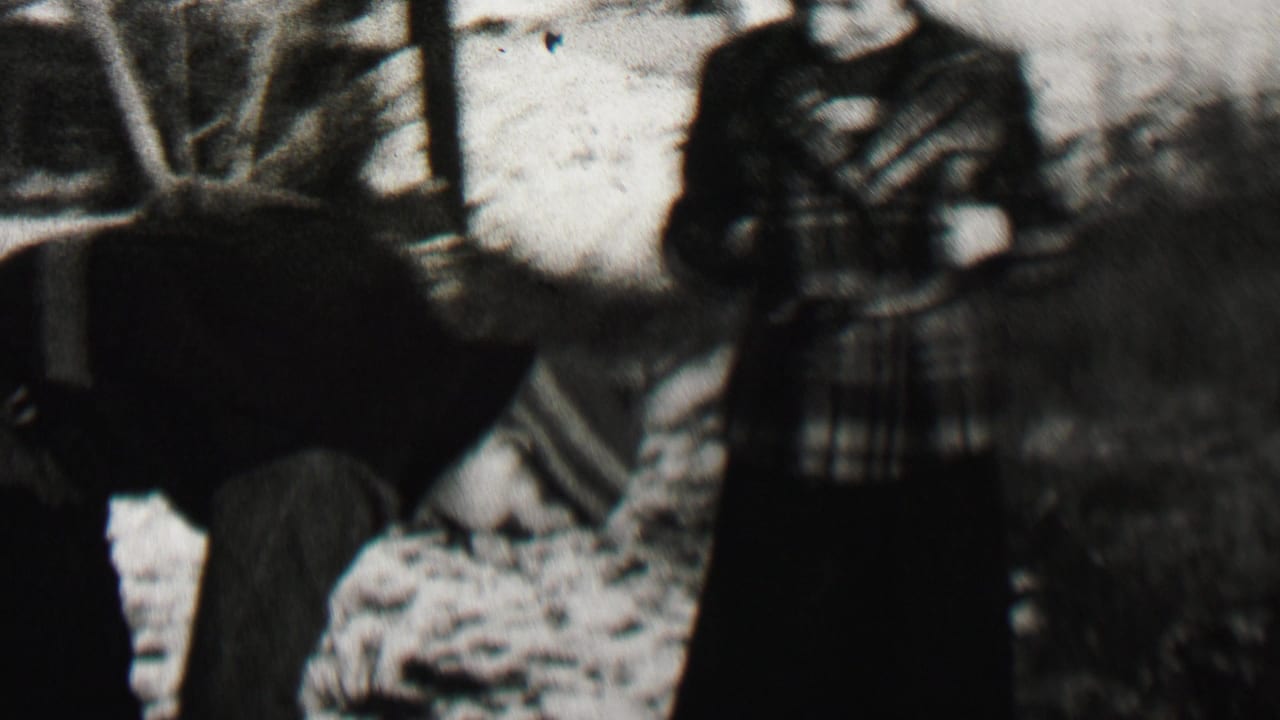 Bundled person bends over snow beside a standing woman in a plaid coat.