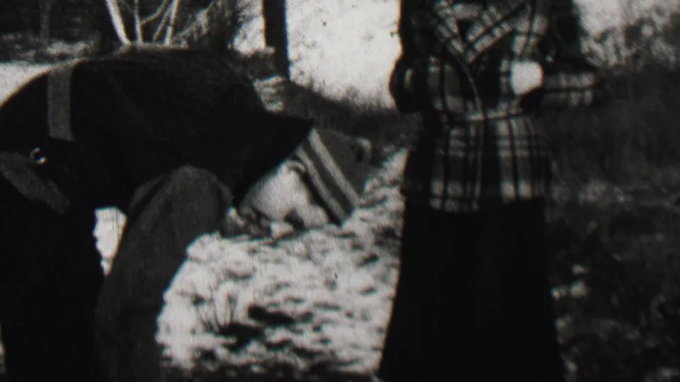 A person in a dark winter coat bends beside the standing woman in plaid, with snow, shrubs, bare trees, and a wall-like background visible in the 1937 Beachwood scene.