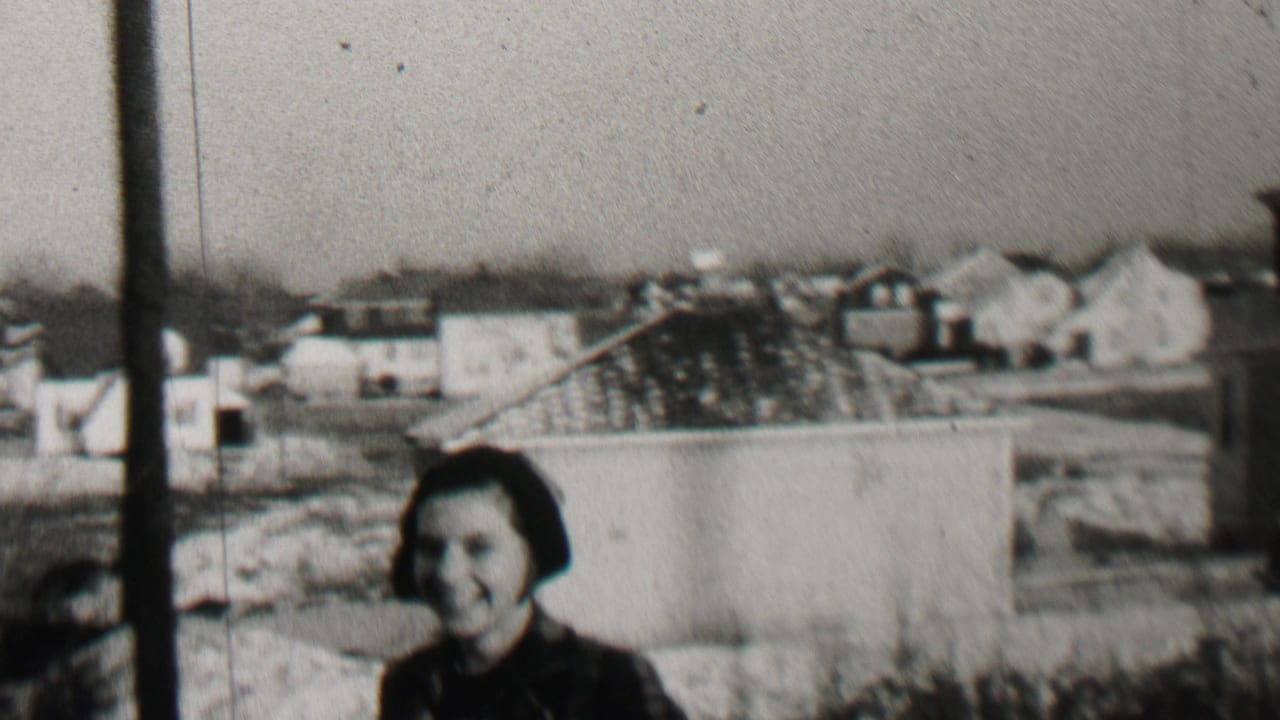 Three children in winter clothing stand and smile outdoors in Beachwood, New Jersey, with residential buildings and rooftops visible behind them in the 1937 home-movie frame.