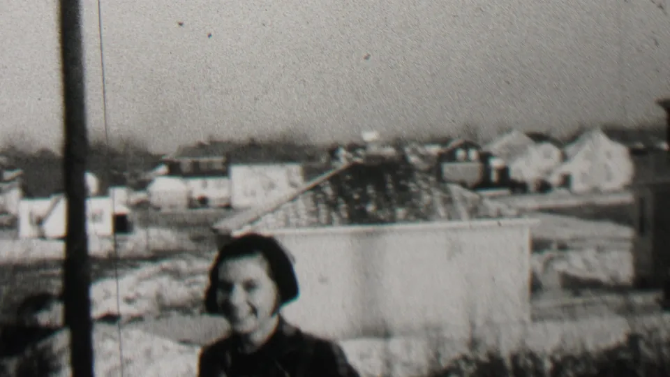Three children in winter clothing stand and smile outdoors in Beachwood, New Jersey, with residential buildings and rooftops visible behind them in the 1937 home-movie frame.