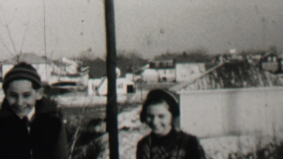 Two bundled children, one facing the camera and one turned partly sideways, stand near a vertical pole in a Beachwood residential yard with pale winter ground and houses behind them.