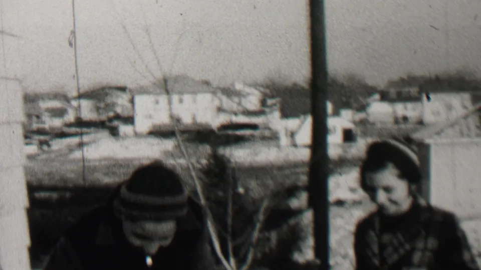 One bundled child bends forward near bare shrubs while another child stands nearby, with houses and open winter ground visible in the Beachwood, New Jersey home-movie frame.