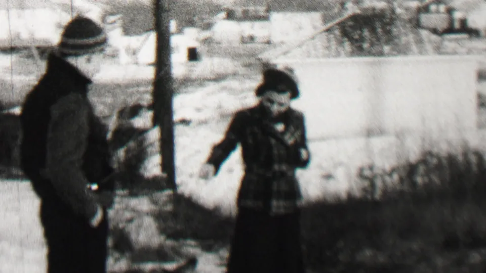 A bundled child stands at right in a patterned coat while another child faces away near a vertical pole, with low houses and winter ground in the background.