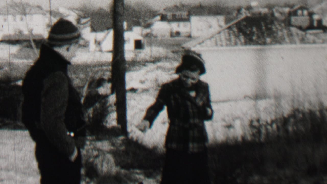 In Beachwood, New Jersey home-movie footage from 1937, an adult man in a dark coat and knit cap stands near an adult woman in a plaid coat and hat beside winter ground and distant houses.