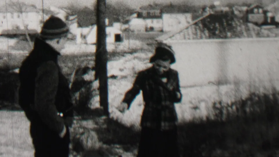 In Beachwood, New Jersey home-movie footage from 1937, an adult man in a dark coat and knit cap stands near an adult woman in a plaid coat and hat beside winter ground and distant houses.