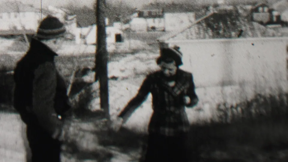 A man in a dark coat and a woman in a plaid coat stand near a utility pole with residential buildings and winter ground behind them in the Beachwood, New Jersey footage.