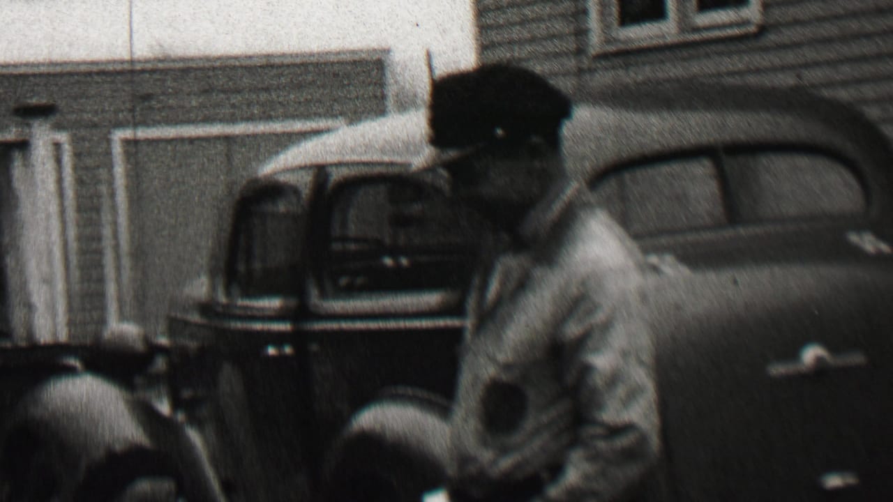 Man in cap and light jacket walking beside parked vintage cars in front of a house.