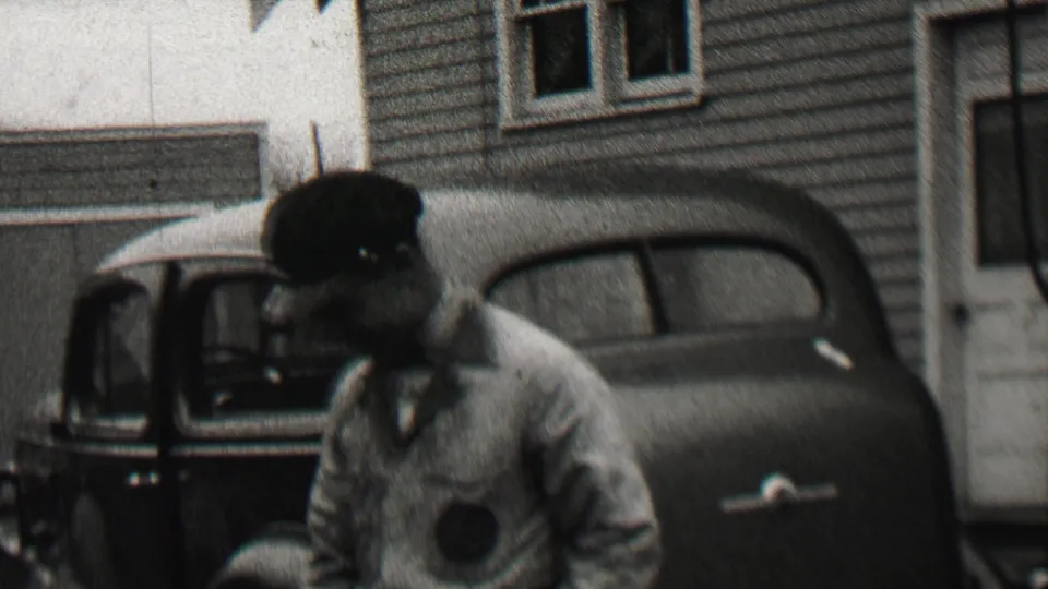 A man in a cap stands near the foreground while a smiling woman appears from inside or beside a parked car in the supplied 1937 Beachwood home-movie scene.