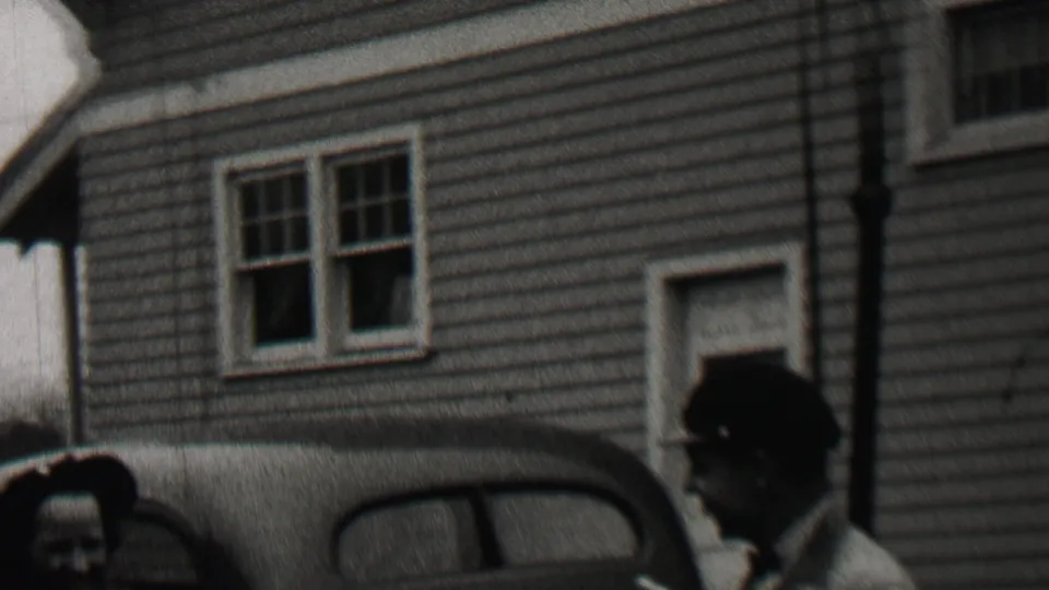 Two adults are partly framed near a parked rounded automobile outside a clapboard house in the Beachwood, New Jersey home-movie scene.