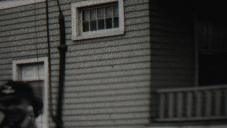 A partially visible adult appears near the edge of the frame as the camera catches the house wall, small window, downspout, and porch railing.