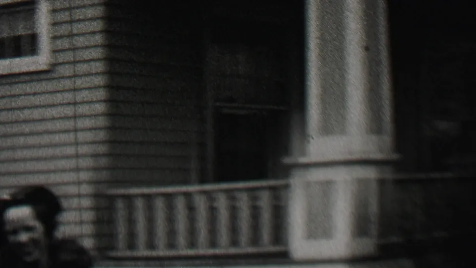 A woman is partially visible near a porch railing and house entrance, with clapboard siding and a doorway filling the residential background.