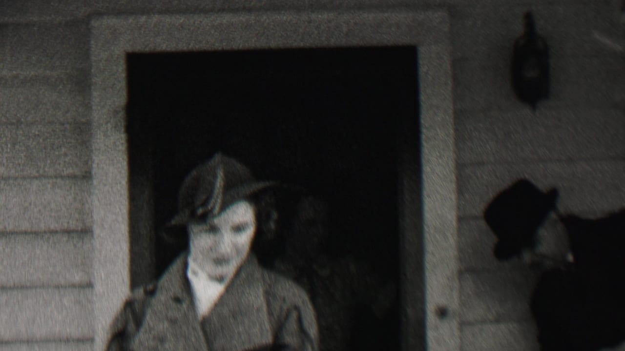 In supplied 1937 Beachwood, New Jersey home-movie footage, a woman in a hat and coat stands by a clapboard house doorway with another hatted adult partly visible behind her.