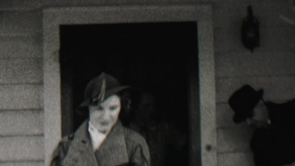 In supplied 1937 Beachwood, New Jersey home-movie footage, a woman in a hat and coat stands by a clapboard house doorway with another hatted adult partly visible behind her.