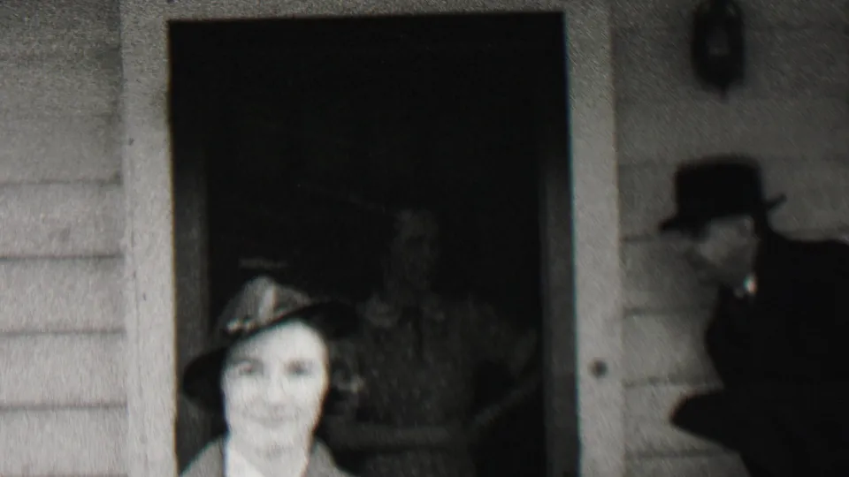 A woman in a small hat faces the camera outside the Beachwood house, with dark doorways, clapboard siding, and partially visible adults in hats around her.
