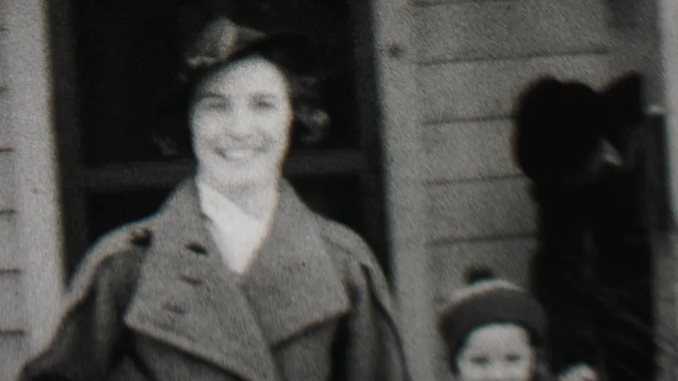 A smiling woman in a hat and overcoat stands close to the camera outside the clapboard house in the supplied 1937 Beachwood home-movie scene.