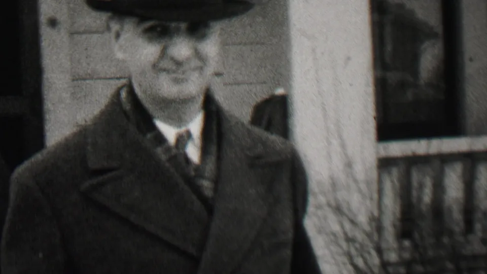 A man in a suit, tie, overcoat, and brimmed hat smiles near a porch post, with a clapboard house exterior and partial figures behind him.