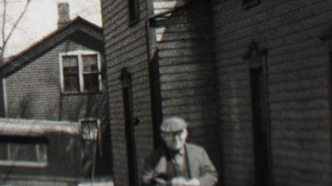 An older man in a flat cap and suit stands or walks near clapboard residential buildings in the supplied Beachwood, New Jersey 1937 footage.
