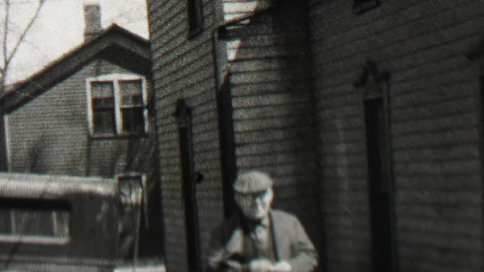 An older man in a flat cap and suit stands or walks near clapboard residential buildings in the supplied Beachwood, New Jersey 1937 footage.