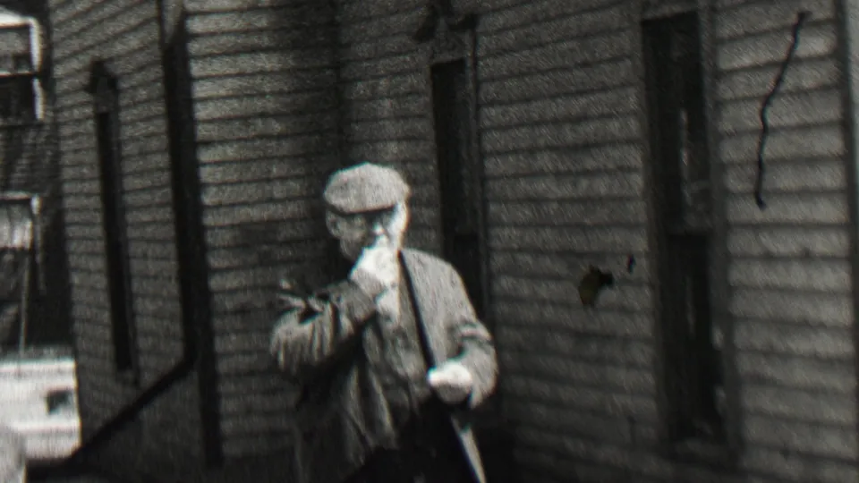 The man in a flat cap appears to eat or touch his mouth while standing near a clapboard house and a parked early automobile in the 1937 Beachwood home movie.