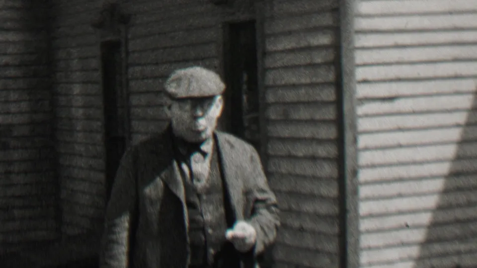 An older man in a suit and flat cap walks beside a clapboard residential building, holding a small object in the 1937 Beachwood, New Jersey footage.
