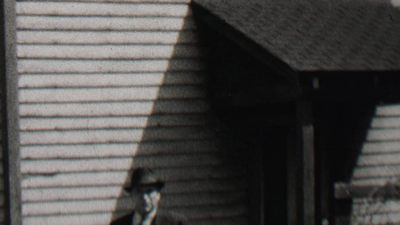 In 1937 Beachwood, New Jersey home-movie footage, a suited man in a fedora walks near a clapboard house while another smaller figure is partly visible at the left edge.