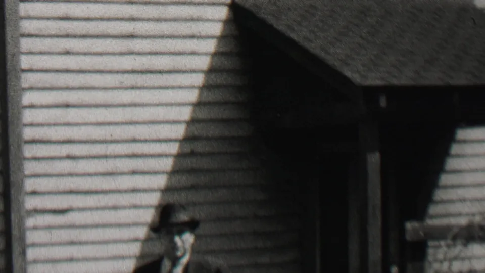 In 1937 Beachwood, New Jersey home-movie footage, a suited man in a fedora walks near a clapboard house while another smaller figure is partly visible at the left edge.