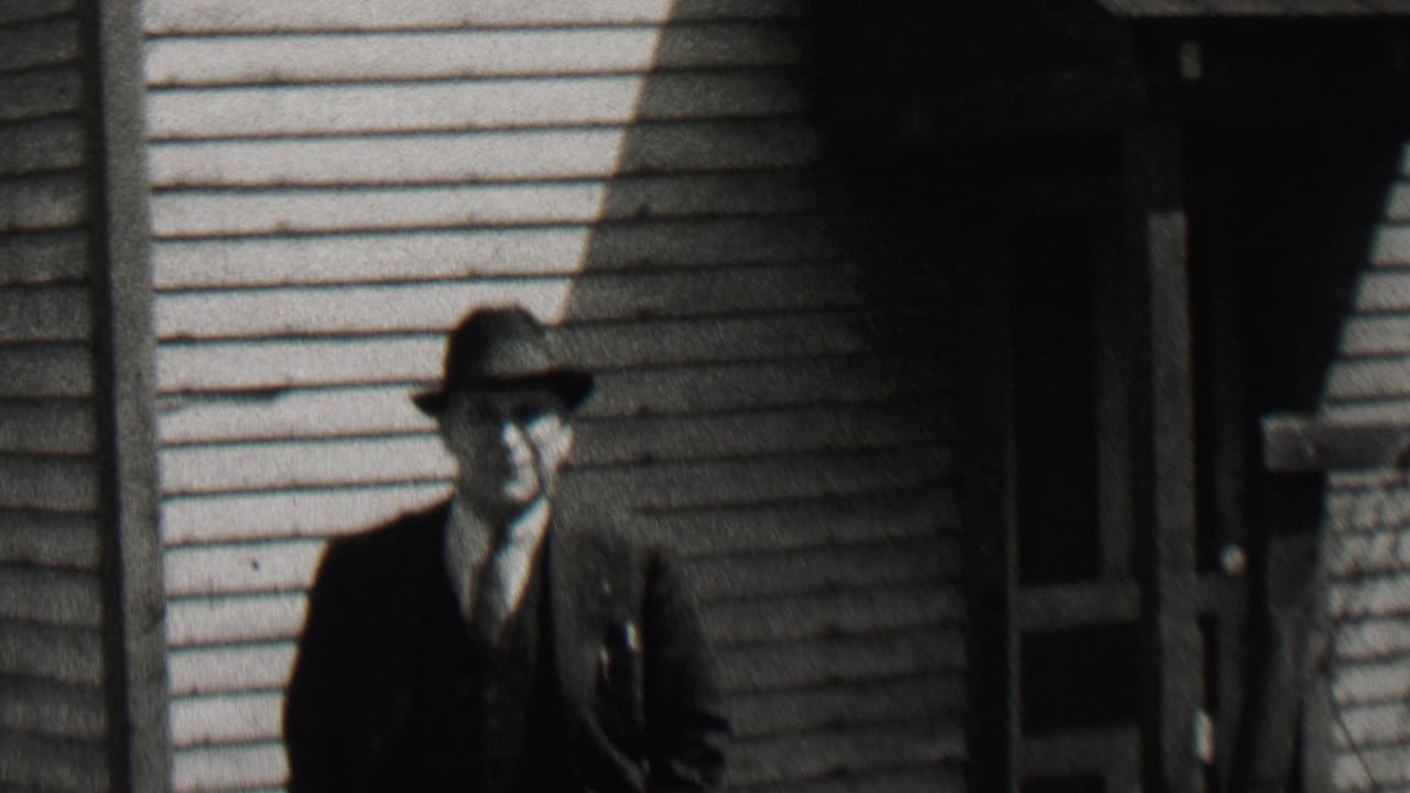 A man in a dark suit, vest, tie, and fedora approaches the camera outside a light-sided house in the Beachwood, New Jersey 1937 home-movie clip.