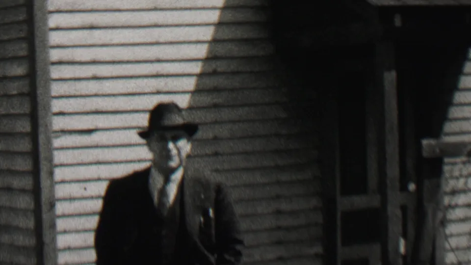 A man in a dark suit, vest, tie, and fedora approaches the camera outside a light-sided house in the Beachwood, New Jersey 1937 home-movie clip.