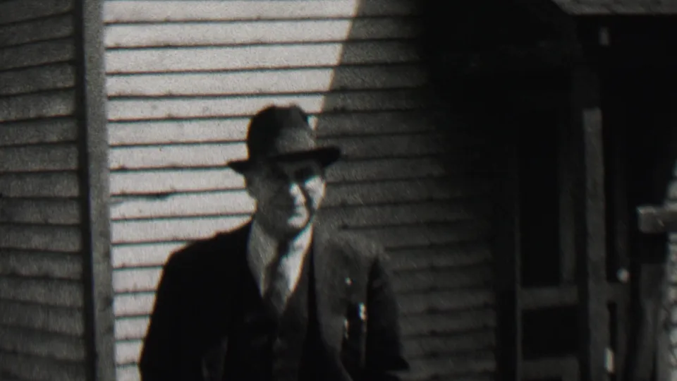 The suited man in a fedora is closer to the camera, smiling slightly as roof shadows fall across the light siding behind him.