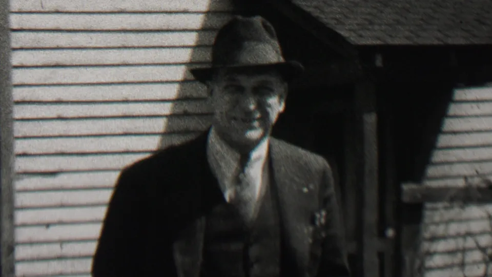 A close view shows the man smiling in a fedora, dark suit, vest, shirt, and tie against a clapboard house exterior in the 1937 Beachwood footage.