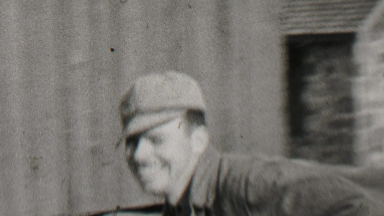 In 1937 Beachwood, New Jersey home-movie footage, a blurred man in a cap passes close to camera while two adult men stand smiling outside a clapboard house.