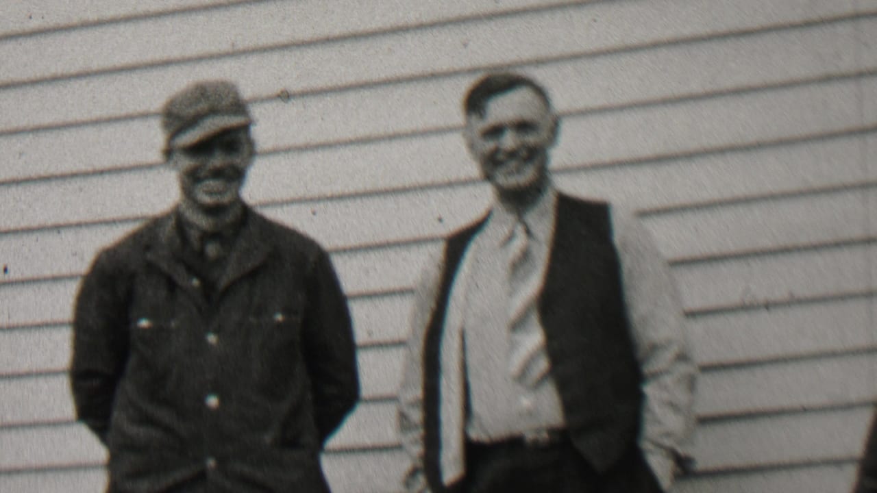 Two smiling men stand beside a house with horizontal siding.