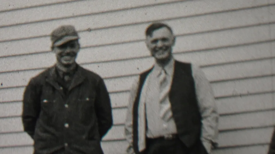 Two adult men smile toward the camera outside a horizontally sided house in Beachwood, New Jersey, one wearing a cap and work jacket and the other a vest and tie.