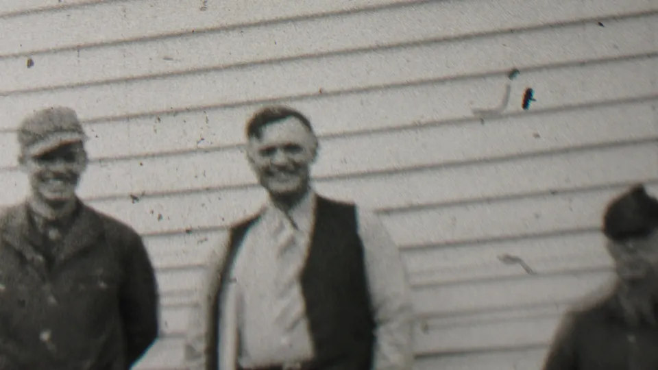 Two men remain posed near the clapboard house as a partially visible person bends or moves in the yard during the 1937 Beachwood home-movie scene.