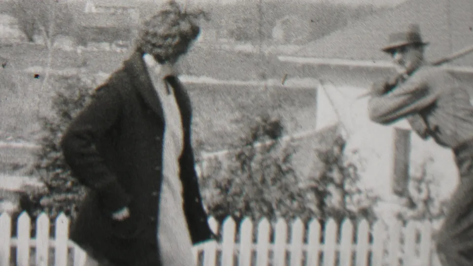 A woman in a long coat stands in a backyard near a white picket fence as a man in a hat swings a golf club in the 1937 Beachwood, New Jersey footage.