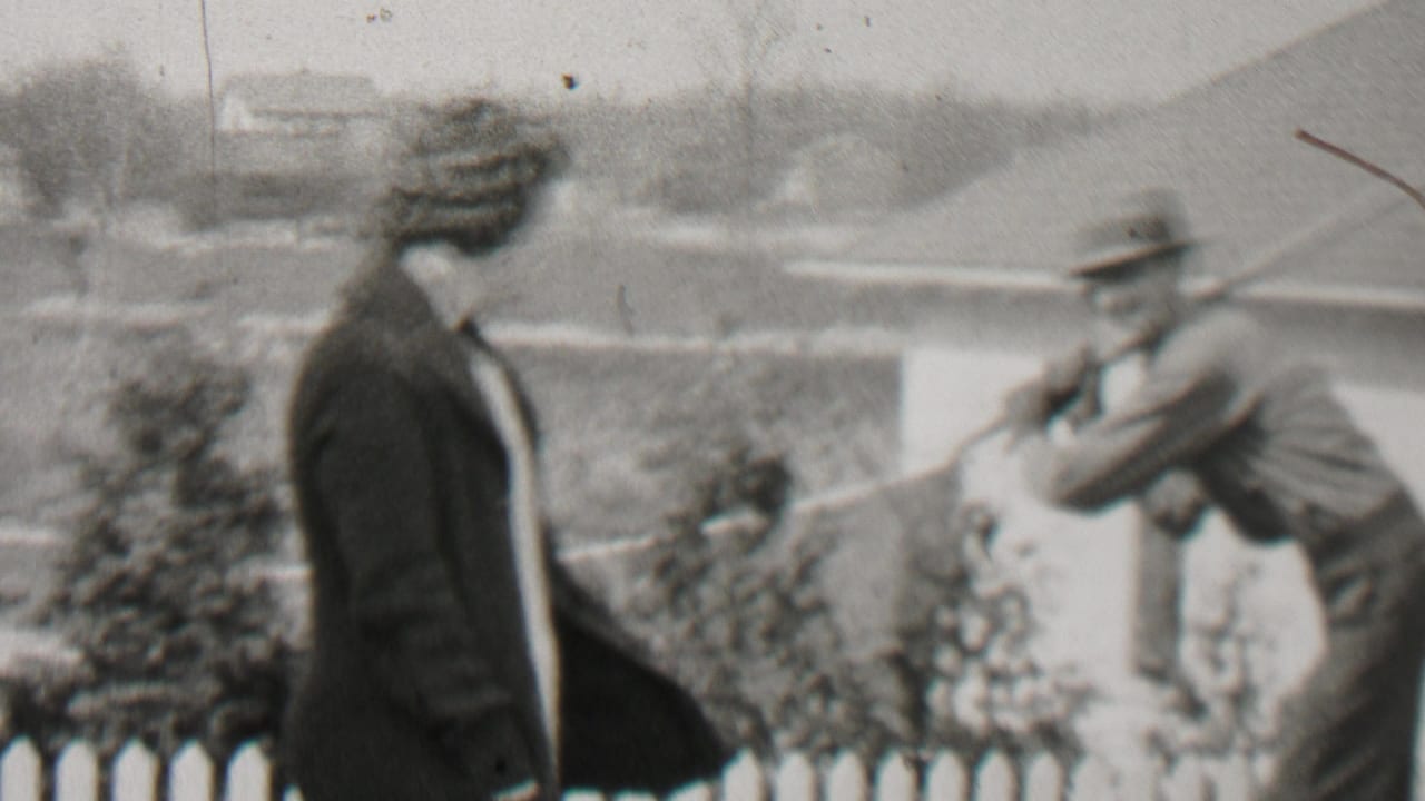 In the 1937 Beachwood, New Jersey home-movie frame, a woman in a dark coat stands by a white fence as a man in a brimmed hat holds a rake nearby.