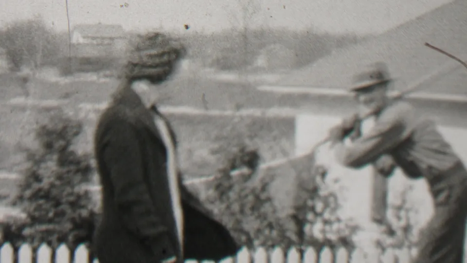 In the 1937 Beachwood, New Jersey home-movie frame, a woman in a dark coat stands by a white fence as a man in a brimmed hat holds a rake nearby.