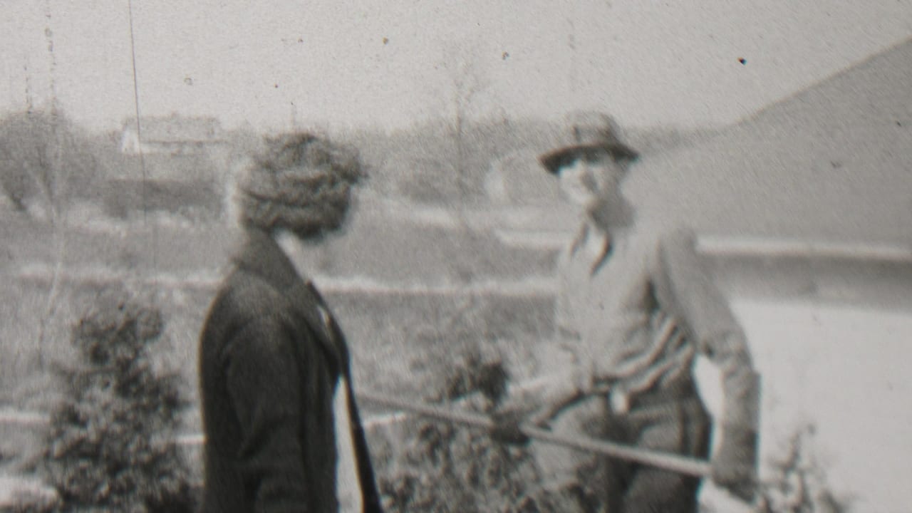 Man in a hat raises a rake while a woman in a dark coat looks on beside a fence.