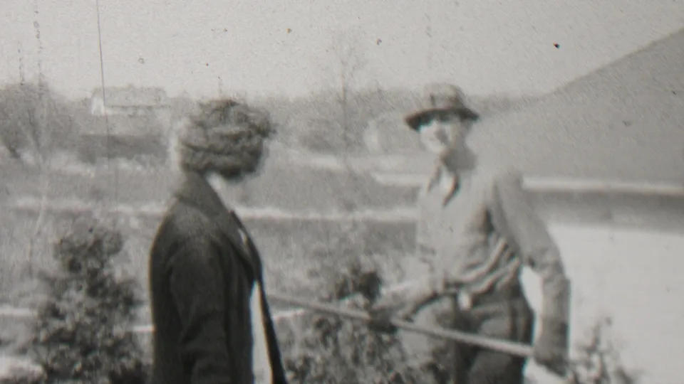 A man in work clothes and a brimmed hat raises a garden rake in a playful stance while a woman in a dark coat watches in the Beachwood yard scene.