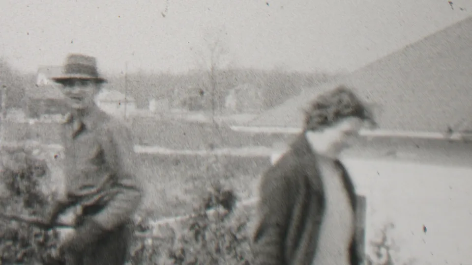 The man with the rake stands at left while the woman bends forward near the fence, with a roofline and shrubs behind them in the 1937 home-movie frame.