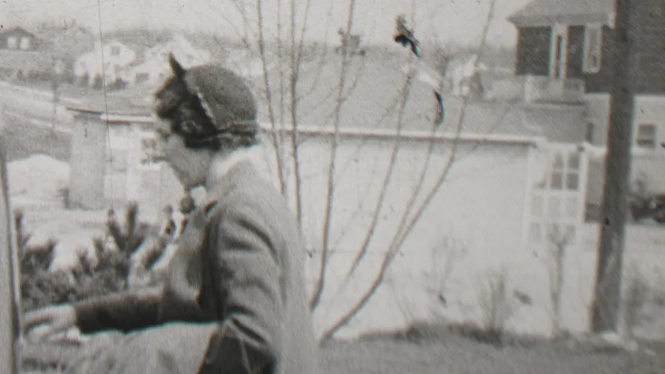 A woman in a coat turns or gestures near a bare young tree, a white fence, and neighboring houses in the Beachwood, New Jersey home-movie scene.