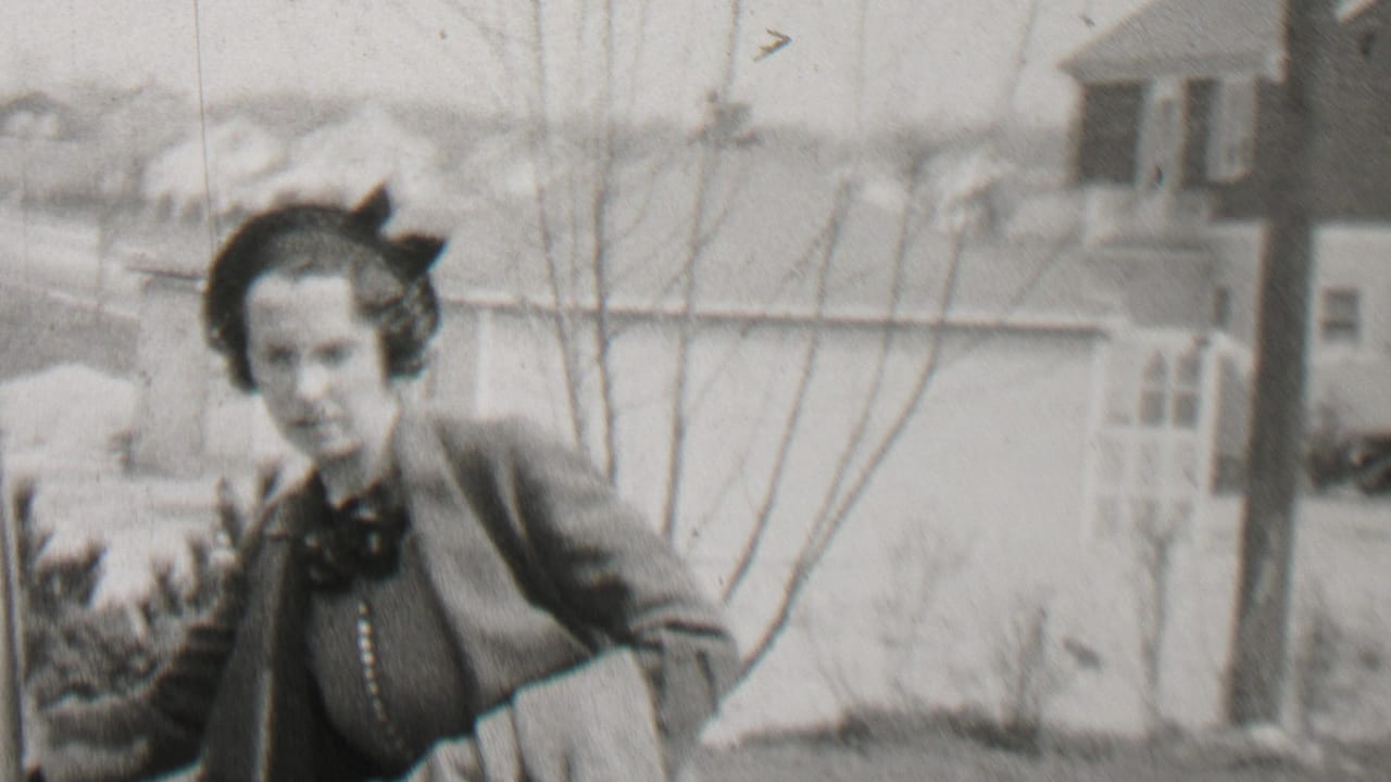 In 1937 Beachwood, New Jersey, a woman wearing glasses, a dark hat, and a dark coat stands close to a classic automobile with suburban houses and bare trees behind her.