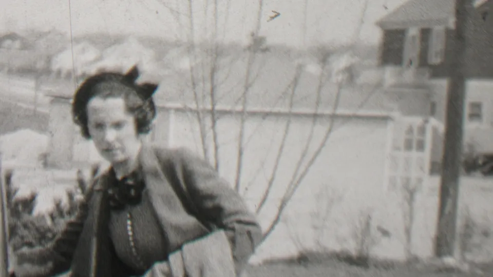 In 1937 Beachwood, New Jersey, a woman wearing glasses, a dark hat, and a dark coat stands close to a classic automobile with suburban houses and bare trees behind her.