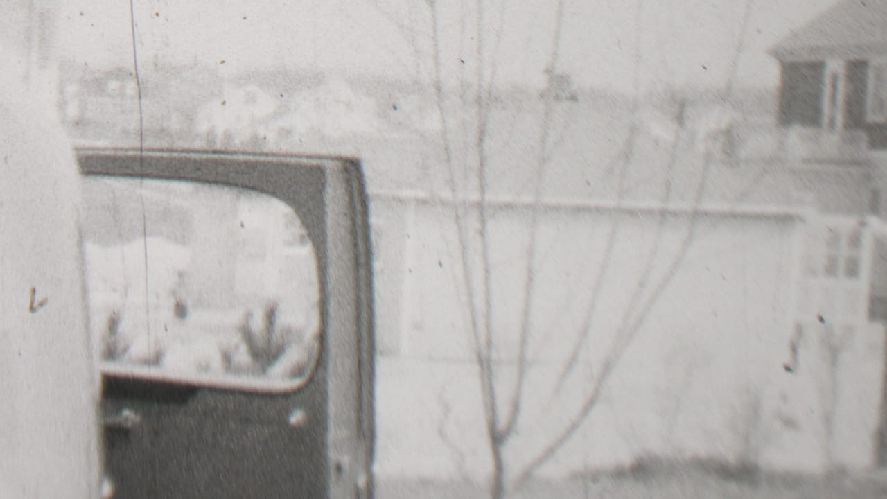 Soft view of a light-colored building, bare tree branches, and part of a vehicle or vehicle body at the left.