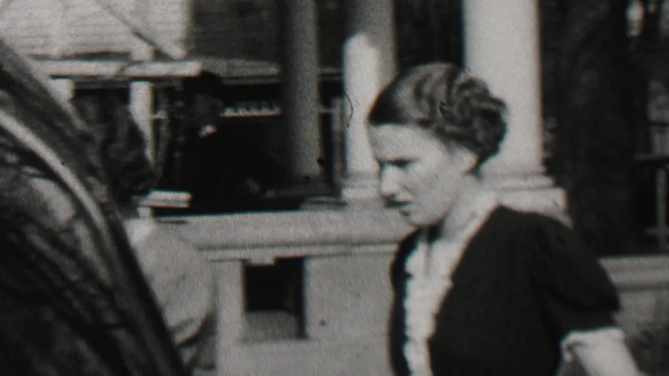 Beachwood, New Jersey 1937 home-movie frame showing an adult woman in a dark dress walking near porch columns and residential houses, with another person partly cropped at the edge.