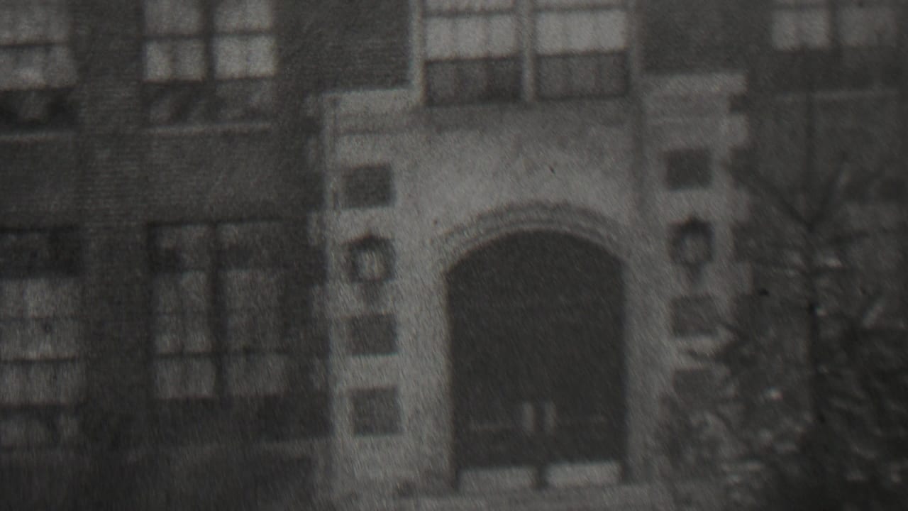 1937 Beachwood, New Jersey 8mm frame showing a soft, grainy exterior view of a brick building with arched doorway openings, multi-paned windows, and winter branches in front.