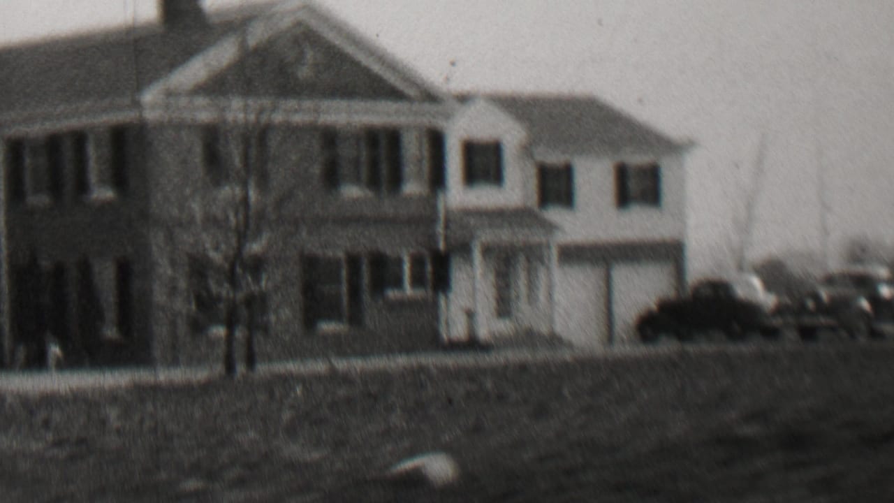 Two-story house with a columned porch, bare trees, lawn, and parked automobiles.